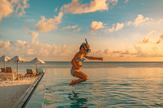 Happy child toddler jumping into infinity swimming pool at sunset as travel lifestyle portrait. Cute girl with blond hair snorkel googles jumps into water splashes water drops. Family summer vacation