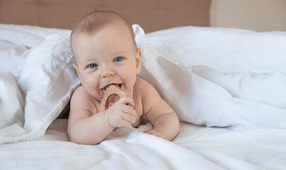 Cute happy baby with wooden teether in hands laying on tummy in bed under white sheets. Little child. Pure emotions. Childhood.