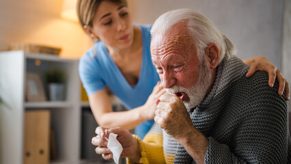 Cold exhausted senior man with flu wrapped in a warm blanket blowing his nose with a tissue in the livingroom. Nurse take care of him.  - obrazy, fototapety, plakaty