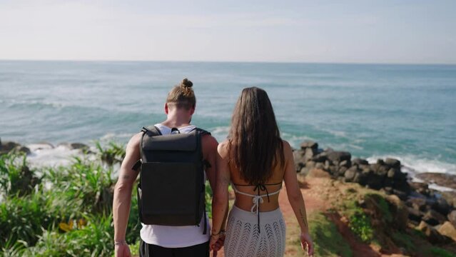 Biracial tourist couple walking towards edge of scenic hill covered with exotic plants above breathtaking view of ocean backview shot. Young adults approach edge of picturescue rock shot from behind