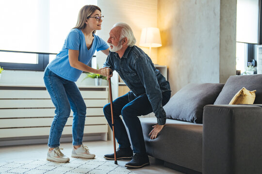 Shot Of A Female Nurse Helping Her Elderly Male Patient. Smiling Senior Man With Female Healthcare Worker. Home Carer Supporting Old Man To Stand Up From The Chair At Care Home.