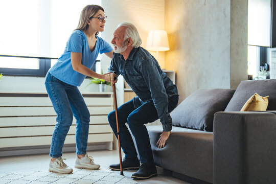 Shot Of A Female Nurse Helping Her Elderly Male Patient. Smiling Senior Man With Female Healthcare Worker. Home Carer Supporting Old Man To Stand Up From The Chair At Care Home.