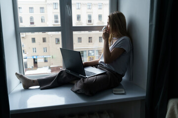 A young woman sits on a windowsill, drinks coffee, works at a laptop, looks out the window.