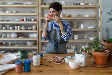 Take a break from work. Creative artisan craftswoman wearing apron drinking morning coffee from handmade clay mug and eating croissant while working in her own cozy pottery studio. Small art business