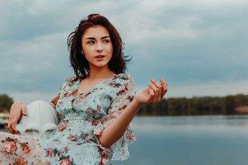 Сharming pretty woman dressed flowery dress standing on the pier near river lake moody cloudy weather in early spring nature. Fashion, girl model with black hair
