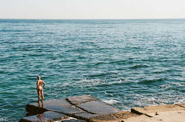 Fototapeta na wymiar Film photograph taken at the beach in Odesa, Ukraine. Elder woman standing by the sea under hot August sun. Retro summer vibes.