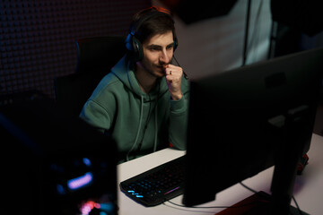 Concentrated male cybersport gamer wearing headset and playing video game having battle in dark room illuminated by neon lights