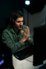High angle of young worried male cybersport gamer sitting at table with prayer hands while playing...