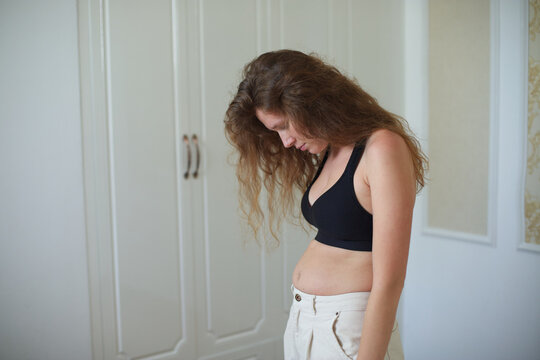 Unhappy sad upset young woman with fat on her stomach, girl worried about body fat touching her belly standing at home near closet