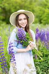 Beautiful young girl in a white dress, straw hat with a bouquet of violet flowers in her hands and picnic basket. Pretty woman in summer in the blooming field holding a bunch of purple lupin
