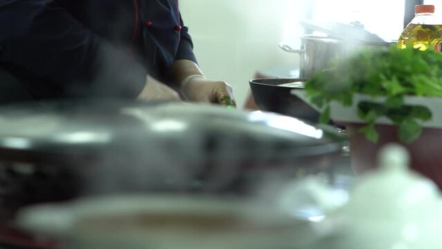 The cook is chopping herbs next to the boiling cauldrons in the kitchen. Steam rises from boiling pots in the kitchen