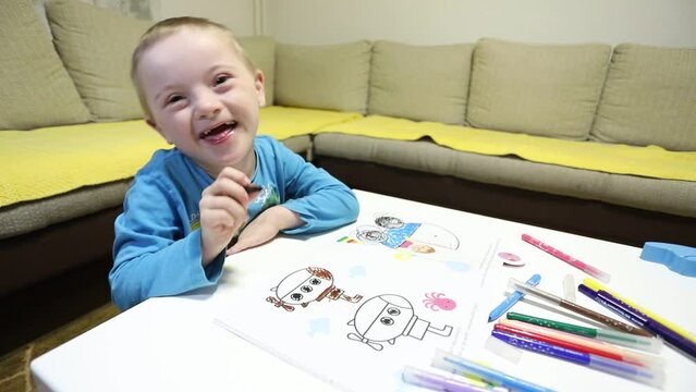 Cute young boy laughs while working on his drawing. People with Down Syndrome. Inclusion, education, home atmosphere. Kids with disability. 21 of March.