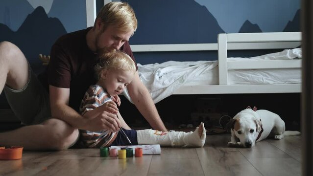 father and son draw with paints on a plaster splint sitting in the children's room