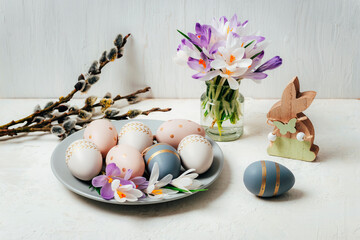 Gray plate with painted Easter eggs and wooden toy bunny, crocus flowers on white table. Easter...
