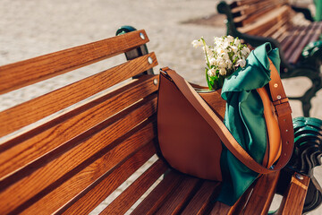 Close up of female leather handbag with tied scarf on bench. Brown purse with bouquet of white...