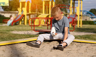 crying child with broken limb arm sits outdoors and looking how kid girl is playing on playground....