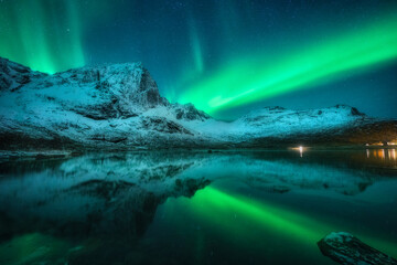 Fototapeta na wymiar Northern lights over the snowy mountains, sea, reflection in water at night in Lofoten, Norway. Aurora borealis and snow covered rocks. Winter landscape with polar lights, sky with stars and fjord
