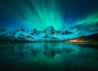 Fototapeta na wymiar Northern lights over the snowy mountains, frozen sea, reflection in water at night in Lofoten, Norway. Aurora borealis and snow covered rocks. Winter landscape with polar lights, starry sky and fjord