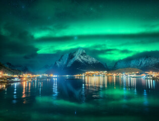 Northern lights in Reine, Lofoten Islands, Norway. Starry sky and Aurora borealis, snowy mountains, sea coast, houses, rocks, reflection in water, city lights at winter night. Bright polar lights
