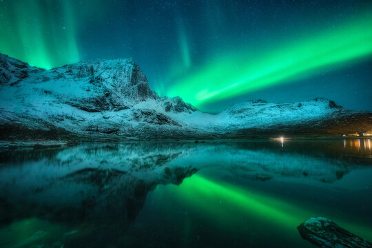 Northern lights over the snowy mountains, sea, reflection in water at night in Lofoten, Norway. Aurora borealis and snow covered rocks. Winter landscape with polar lights, sky with stars and fjord © den-belitsky