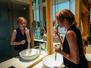 Attractive mature woman is standing in a bathroom putting on makeup