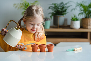 Adorable 3 years old caucasian blonde little girl is watering new plants in the pots at home....