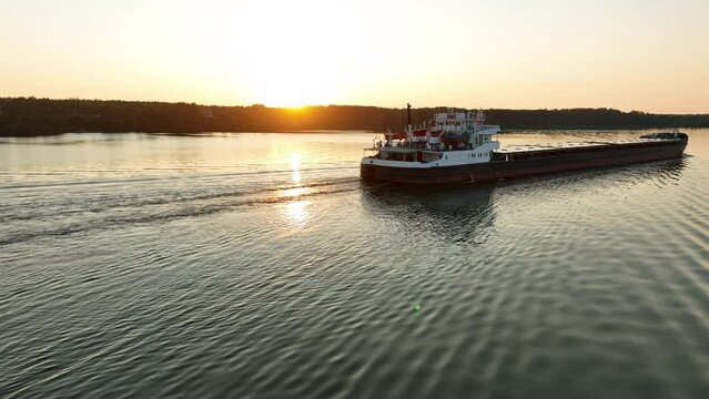 Aerial view. A dry cargo ship sails on the river at sunset. Transportation of goods by water transport. Delivery of raw materials and goods by river. 