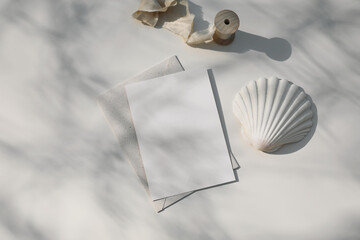 Summer marine neutral stationery, desktop mock-up scene. Blank greeting card, beige envelope. Beige table background in sunlight. Tree branches shadow overlay. Scallop shell and silk ribbon. Flat lay.