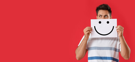 Handsome young man holding paper with drawn smiling emoticon on red background with space for text