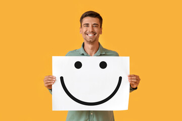Smiling young man holding paper with happy emoticon on yellow background