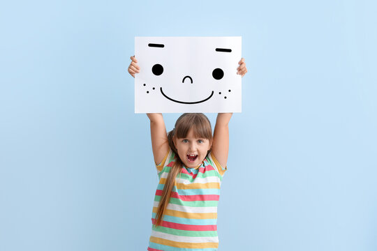 Happy little girl holding paper with smiling emoticon on light blue background