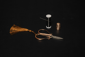 A vintage style copper crafted scissors and thimble along with a spool of shiny black thread on a black surface