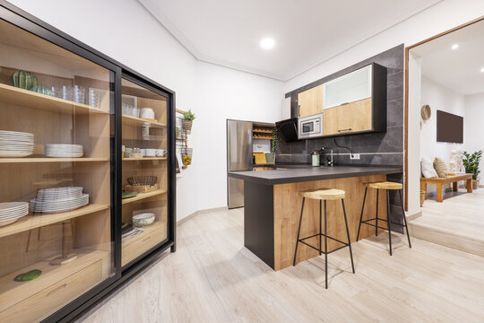 Modern Style Kitchen With Wooden Furniture Combined With Black, A Counter With Stools And A Sideboard For Dishes With Sliding Glass Doors