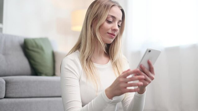 Portrait of beautiful blond woman hold smartphone watching social media with sport bottle take a break after training and looking at camera at home Smiling young woman texting on phone at light room