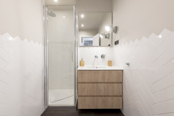 Fototapeta na wymiar Bathroom with white porcelain sink, chrome faucets, frameless mirror, three-drawer wooden wall cabinet, and tempered glass shower stall door