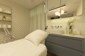 Aesthetic treatment room with a relaxation table, creams and appliances to apply massages and cleansing together with a cabinet with gray drawers, mirrors and a porcelain sink