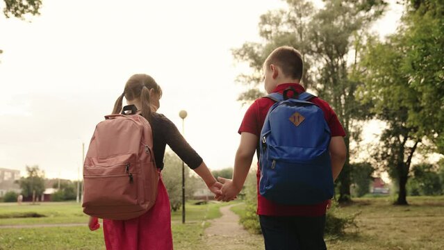 boy girl child holding hands go school sun glare. child with school backpacks his back at sunset. friends child boy girl go lesson park rays sunlight. happy childhood schoolchildren with school bags
