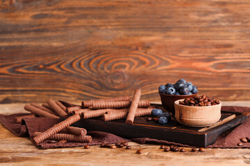 Board with delicious chocolate wafer rolls, blueberries and coffee beans on brown wooden table