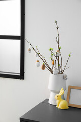 Vase with tree branches, Easter eggs and rabbit on table near grey wall