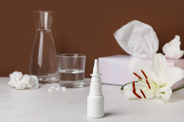 Fototapeta na wymiar Nasal drops with flower, glass of water and tissues on table near brown wall. Seasonal allergy concept