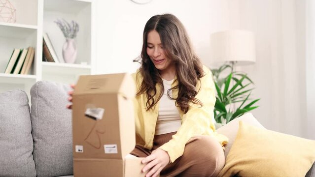 Portrait of young woman receive parcel dissatisfied with shopping in clothes online internet store sitting on sofa at home Upset customer disappointed with purchase order while open carton box indoors