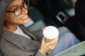 Smiling woman passenger in eyeglasses drinking takeaway coffee in taxi car on the way to work