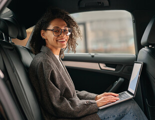 Smiling woman analyst working on laptop sitting car backseat on way to office and looking at camera