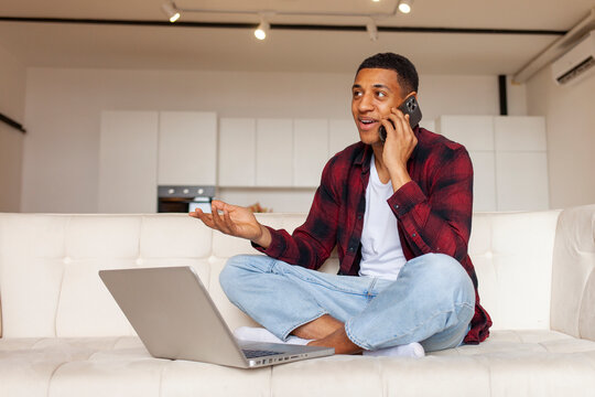 Young African American Guy Sitting At Home On The Couch Using Laptop And Talking On The Phone