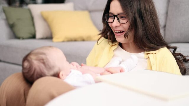 Portrait of pretty young woman mother playing with little baby enjoying time together while writing on blank notebook mom diary at home Charming female try to work or study with newborn child indoors