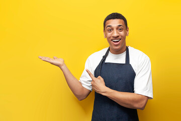 young guy afro american waiter in apron advertises copy space on yellow isolated background,...