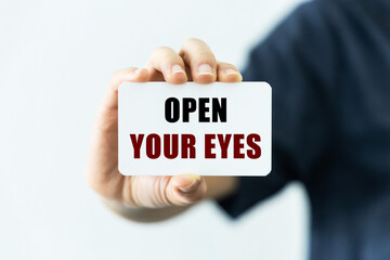Open your eyes text on blank business card being held by a woman's hand with blurred background. Business concept about opening your eyes.