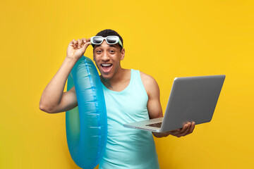 cheerful african american guy in sunglasses with inflatable swim ring uses laptop on yellow...