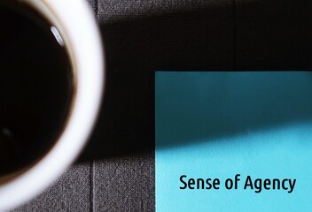 Blurred coffee mug with blue paper note written SENSE OF AGENCY, sensation of control over our...