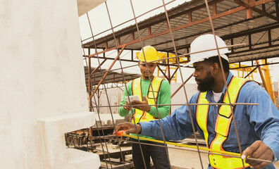 African American foreman uses tape measure to check the spacing rebar used to lay precast concrete walls to a solid and stable standard. Asian male workers record their work on a tablet.